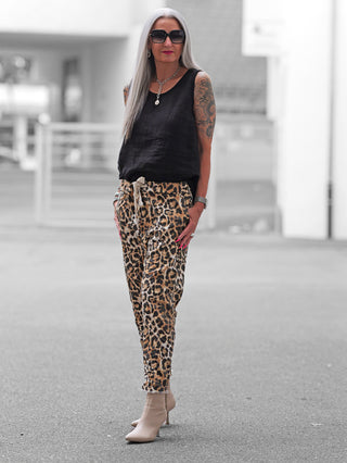Person trägt OFF#DLY Leo Joggpant Ponzano mit auffälligem Leopardenmuster und bequemem Schnitt. Kombiniert mit schwarzem Top, beigen Stiefeletten und silbernen Accessoires für einen stilvollen, urbanen Look.