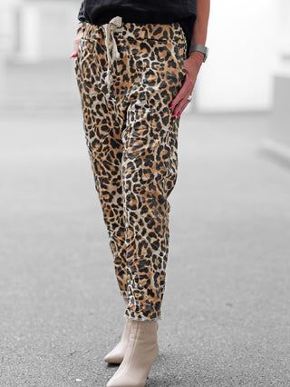 Person in leopard-print OFF#DLY Leo Joggpant Ponzano mit Kordelzug, kombiniert mit beigefarbenen spitzen Ankle Boots. Eine Hand in der Tasche, andere mit pinkem Nagellack, Uhr und Ring sichtbar.