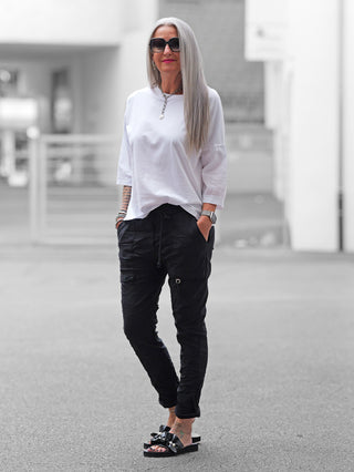 Frau trägt OFF#DLY Pant Iseo black, eine schwarze, lässige Joggpant mit Gummibund und Kordelzug, kombiniert mit weißem Oversized-Shirt und schwarzen Sandalen, verkörpert urbanen Chic.