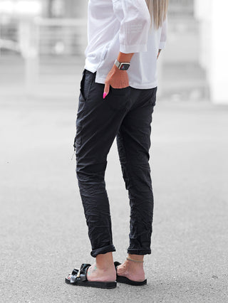 OFF#DLY Pant Iseo black: Schwarze Joggerhose mit leichtem Crinkle-Look, elastischem Bund und umgeschlagenen Knöcheln, kombiniert mit weißen Oberteilen und Sandalen, betont durch eine schlanke femininer Silhouette.