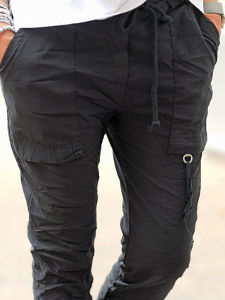 Die OFF#DLY Pant Iseo black zeigt einen urbanen Stil mit sportlicher Note, featuring einen Kordelzugbund, seitliche Taschen und Cargo-Details aus leichtem, crinkle-nylonartigem Stoff. Silberschmuck und Uhr ergänzen den Look.