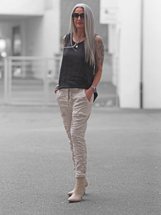 Frau trägt OFF#DLY Pant Iseo beige: locker sitzende, leicht geraffte Joggpant mit Kordelzug und seitlichen Taschen. Kombiniert mit schwarzen Oberteilen und cremefarbenen Heels für urbanen Chic.