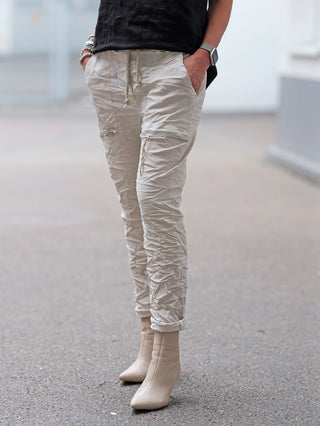 OFF#DLY Pant Iseo beige: Person trägt beige, leicht geknitterte Cargo-Jogger mit Kordelzug und Seitentaschen, kombiniert mit spitzen Stiefeletten. Hände in Taschen, Armbänder und Smartwatch sichtbar.