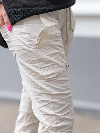 Nahaufnahme der OFF#DLY Pant Iseo beige, betont die leicht verknitterte Baumwollstruktur mit Kordelzug und Seitendetail. Eine Hand mit pinken Nägeln steckt lässig in der Hosentasche.