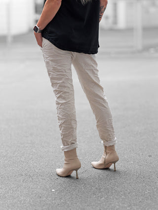 Person trägt OFF#DLY Pant Iseo beige, leicht gekrempelt, mit Gummibund und Crinkle-Optik, kombiniert mit beigen Kitten-Heel-Stiefeletten und schwarzem Oberteil. Sichtbar sind Smartwatch und kleines Tattoo am Unterarm.