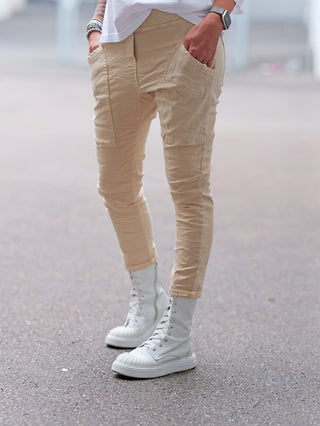 OFF#DLY Joggpant Vellano beige: Slim-fit Cargo-Hose mit Taschen und gerafften Beinabschlüssen, getragen mit weißen Stiefeln. Hände in den Taschen, Armbanduhr und Armbänder sichtbar.