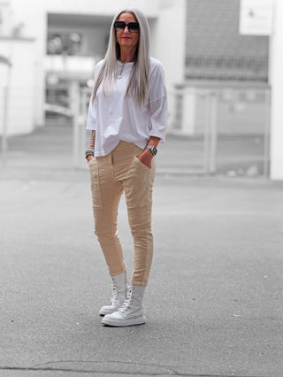 Frau trägt OFF#DLY Joggpant Vellano beige, locker sitzend mit Cargotaschen und elastischem Bund, kombiniert mit weißem T-Shirt und weißen Stiefeletten, im urbanen Hintergrund.