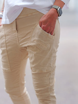 OFF#DLY Joggpant Vellano beige, getragen mit weißem Oberteil, zeigt eine große, schräg platzierte Seitentasche und eine moderne Smartwatch am Handgelenk. Perfekt für sportlich-lässigen Sommerlook.