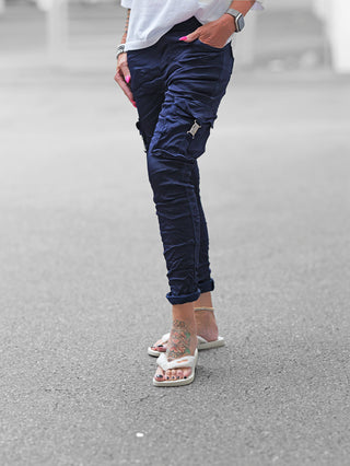 OFF#DLY Pant Otranto darkblue: Schlanke, navyblaue Cargohose mit mehreren Taschen und Metallschnallen, kombiniert mit weißen Flip-Flops. Sichtbare Accessoires: Smartwatch, Armbänder, florales Fußtattoo, rosa lackierte Fingernägel.