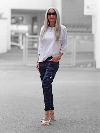 Frau trägt OFF#DLY Pant Otranto darkblue, eine elegante Cargohose mit silbernen Details und elastischem Bund. Kombiniert mit weißem Oberteil und Flip-Flops zeigt sie einen modernen, lässigen Look.