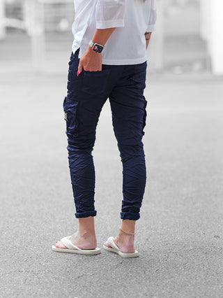 Person trägt OFF#DLY Pant Otranto darkblue mit schlanker Passform und eleganten Cargodetails. Hose leicht gekrempelt, kombiniert mit weißen, flauschigen Sandalen, pinken Nägeln und Smartwatch.