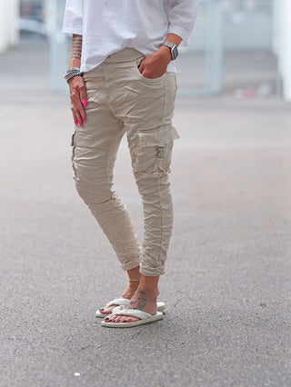 Person trägt OFF#DLY Pant Otranto champagner mit gerollten Bündchen, ergänzt durch weiße, flauschige Sandalen. Sichtbar sind eine florale Fußtattoo, ein Fußkettchen, pinke Fingernägel und eleganter Schmuck.