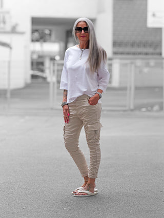 Frau trägt OFF#DLY Pant Otranto champagner, eine beige Cargo-Hose mit Taschen und elastischem Bund, kombiniert mit weißem Top und Sandalen, in urbaner Umgebung. Luxuriös und bequem.