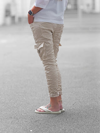 Die OFF#DLY Pant Otranto champagner zeigt sich in hell-beige mit Cargotaschen und silbernen Details. Sie kombiniert Komfort und Eleganz, getragen mit weißen, flauschigen Slippern und einem Smartwatch am Handgelenk.