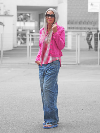 Frau trägt die OFF#DLY Jacke Genua fuchsia in leuchtendem Pink. Die Jeans-Style-Jacke verleiht ihrem lässigen Look mit Baggy-Jeans und Flip-Flops eine urbane Note, während sie vor einem Tor steht.