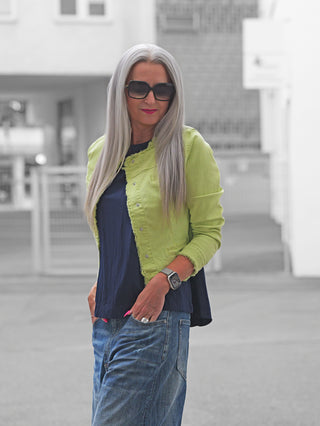 Frau trägt die OFF#DLY Jacke Turin lime, eine figurbetonte, limettengrüne Jacke mit Raw-Edge-Look, über einem dunkelblauen Top. Hände in den Taschen, urbanes Setting.