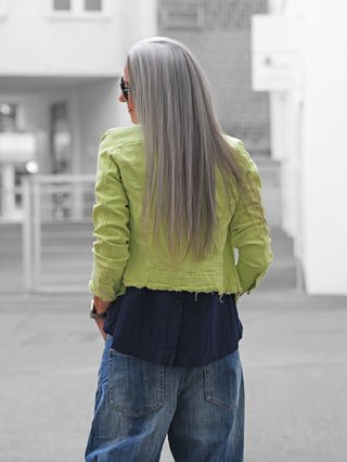Person mit langen grauen Haaren trägt die OFF#DLY Jacke Turin lime, eine figurbetonte, lime-grüne Jacke mit Raw-Edge-Look, über einem dunklen Shirt und blauen Jeans in urbaner Umgebung.