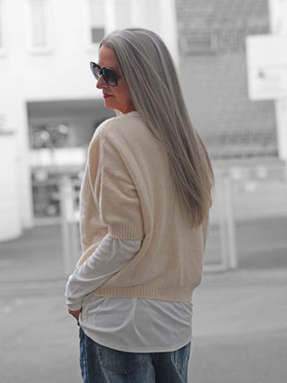 Person im urbanen Umfeld trägt den BSB Cardigan Ivoire, ein kurzärmeliger, cremefarbener Strickcardigan mit Perlmuttknöpfen, über einem weißen Langarmshirt, kombiniert mit lässigen blauen Jeans.