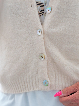 Nahaufnahme des BSB Cardigan Ivoire: Ein weiches, cremefarbenes Strickstück mit schimmernden Perlmuttknöpfen, kombiniert mit einem funkelnden Top und einem Hauch von Animal-Print, getragen von einer Person mit pinken Nägeln.