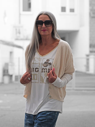 Frau mit langen grauen Haaren trägt den BSB Cardigan Ivoire, ein beige, kurzärmeliges Strickstück mit Perlmuttknöpfen, über einem goldverzierten weißen Top, kombiniert mit blauen Jeans und silbernen Ringen.