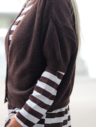 Person trägt BSB Cardigan chocolate: kurzärmeliger, dunkler, flauschiger Cardigan mit Perlmuttknöpfen über gestreiftem Langarmshirt. Teil des Torsos und Armes sichtbar. Ideal für elegante Layering-Looks.