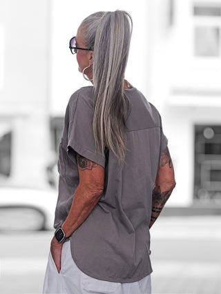 Person mit langen grauen Haaren im hohen Pferdeschwanz trägt ein OFF#DLY T-Shirt Verbania taupe, locker geschnitten mit rundem Saum. Sichtbare Tattoos, silberne Creolen, schwarze Brille, und Smartwatch ergänzen den Look.