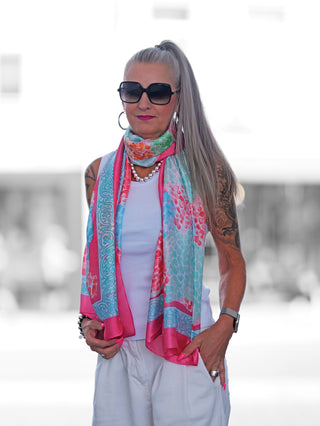 Person trägt den OFF#DLY Schal Mosaik pink in leuchtenden Pink-, Aqua- und Orangetönen, elegant um den Hals drapiert, passend zu einer schlichten weißen Kleidung und auffälligen Accessoires.