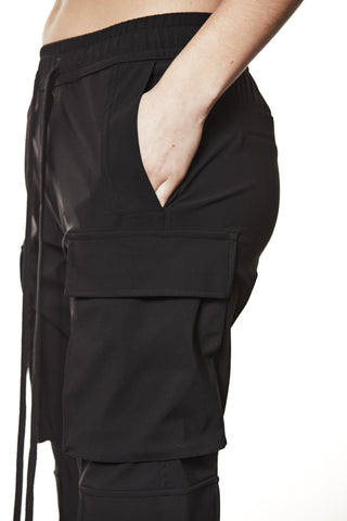 Thom Krom Pant WP 15 black zeigt einen sportlichen Cargo-Look mit elastischem Bund, Fronttasche und großer Cargo-Tasche am Oberschenkel. Der rechte Arm steckt lässig in der Fronttasche.