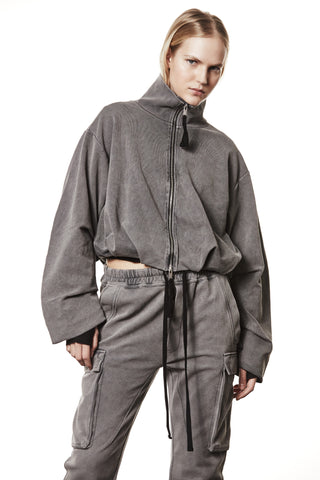 Modell trägt eine Thom Krom Jacke WSJ 494 used grey, oversized mit Stehkragen, Zweiwege-Reißverschluss, überschnittenen Schultern, elastischem Saum und Tunnelzug, kombiniert mit passenden Cargo-Joggern für einen lässigen Athleisure-Look.