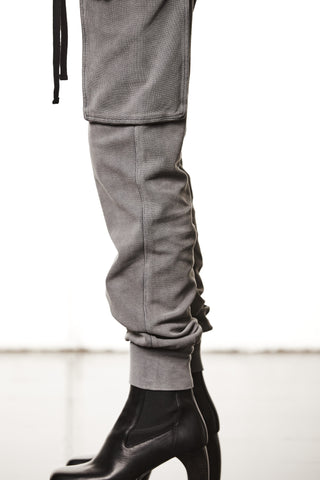 Nahaufnahme eines Beins mit Thom Krom Pant WST 417 in Used Grey, einer grauen Jogginghose mit umgeschlagenem Saum und Klappentasche am Oberschenkel, kombiniert mit schwarzen Lederstiefeletten.