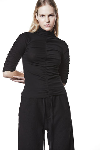Junger Erwachsener trägt das Thom Krom T-Shirt WTS 582 black mit gerüschter Mittelnaht, hohem Kragen und 3/4-Ärmeln. Kombiniert mit schwarzen, locker geschnittenen Hosen für minimalistischen Look.
