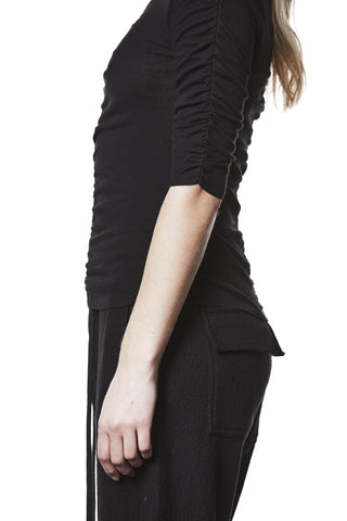 Thom Krom T-Shirt WTS 582 black zeigt einen figurbetonten, schwarzen Longsleeve mit gerafften Ärmeln und asymmetrischen Nähten, getragen zu schwarzen Hosen mit Klappentasche, vor weißem Hintergrund.