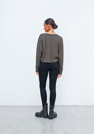 Person von hinten in OUTRYGHT RIB Longsleeve faded black und hochgeschnittenen schwarzen Skinny-Jeans. Kombiniert mit klobigen schwarzen Plattformstiefeln, dezentem Schmuck und tiefem Dutt vor weißer Wand.