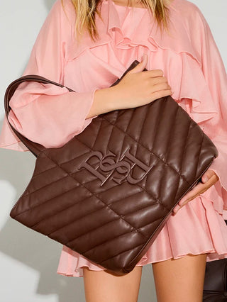 BSB Shopper brown, eine große dunkelbraune Ledertasche mit langen Schulterriemen, diamantgestepptem Muster und erhabenem, ineinandergreifendem Logo vorne, getragen von Person in rüschigem Minikleid.