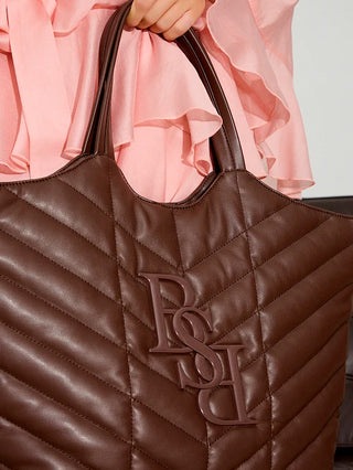 Nahaufnahme einer Person mit einer großen, dunkelbraunen BSB Shopper brown Tasche mit Chevron-Steppmuster und erhabenem Monogramm, gehalten neben einer Hand, die einen pinken Rüschenärmel trägt.