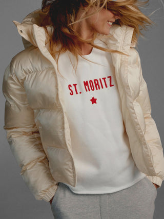 Person in offenem, glänzendem, hellcremefarbenem Pufferjacke, trägt darunter einen Penn&Ink Sweater print ecru/tomato mit rotem ST. MORITZ-Aufdruck und Stern, kombiniert mit hellgrauer Jogginghose.