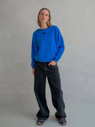 Eine junge Frau trägt den Penn&Ink Sweater print royal/black mit Logo auf der Vorderseite. Kombiniert mit schwarzen Jeans und Trail-Sneakers, vermittelt der Look eine lässige, minimalistische Ausstrahlung.