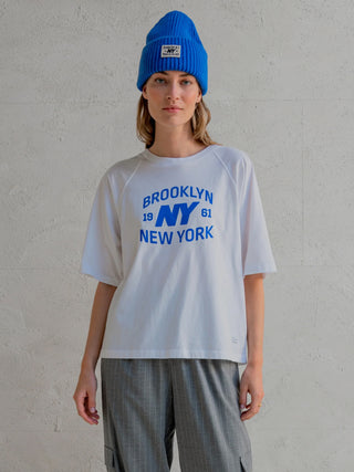 Eine Person trägt das Penn&Ink Tee print white/royalblue mit kurzen Ärmeln und einem Logoprint BROOKLYN NY 1961 NEW YORK auf der Vorderseite, kombiniert mit grauen, gestreiften Hosen.