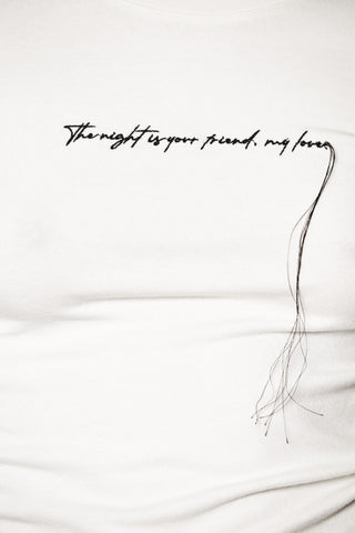 Thom Krom Sweat WTS583 cream zeigt eine Nahaufnahme eines weißen Langarmshirts mit schwarzer Stickerei „The night is your friend, my love“ und losen Fäden auf der Brust.