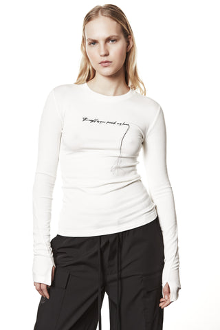 Junge Frau trägt das Thom Krom Sweat WTS583 cream, ein figurbetontes Langarmshirt mit Daumenlöchern und Statement-Stickerei auf der Brust, kombiniert mit lockeren schwarzen Hosen.
