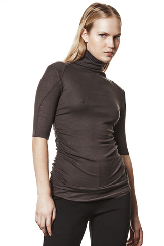 Thom Krom Tee WTS603 night brown: Frau mit langen, blonden Haaren trägt ein figurbetontes, dunkelbraunes Rollkragen-T-Shirt mit Dreiviertelärmeln und seitlichen Raffungen, kombiniert mit schlichten schwarzen Hosen.