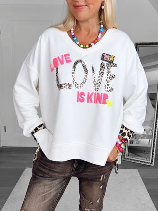 Person in Schlafzimmer trägt den OFF#DLY Sweater LOVE white mit LOVE in Leopardenmuster und LOVE IS KIND in Neonpink. Kombiniert mit Leoparden-Ärmeldetails, dunklen Jeans und buntem Schmuck.