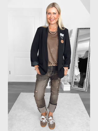 Frau in lässigem Look trägt den OFF#DLY Blazer Bianca black, kombiniert mit Jeans und Sneakers, im Wohnzimmer, mit Accessoires wie Ketten und Gürtel, vor einem Spiegel stehend.