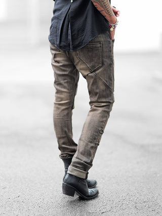 Person trägt OFF#DLY Denim Cecina black Denim: Slim-Fit-Jeans mit rustikalen Nähten und umgeschlagenem Saum, kombiniert mit schwarzen Lederstiefeln. Beaded Armbänder und tätowierter Unterarm ergänzen den urbanen Look.
