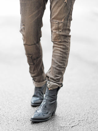 Nahaufnahme von Beinen in OFF#DLY Denim Cecina black Denim, einer urbanen Jeans mit dezenten Rissen und markanten Taschen, getragen mit schwarzen Lederstiefeln auf gepflastertem Untergrund.