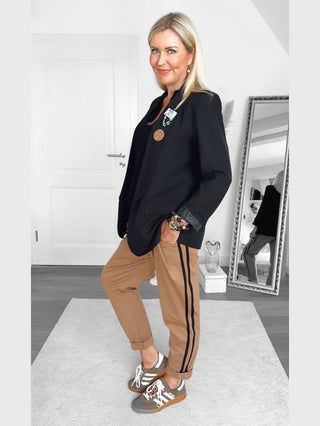 Frau trägt OFF#DLY Pamt Turin camel: schwarze Blazer, schwarze Top, camel-farbene Hose mit schwarzen Streifen. Sneakers in Braun-Grau. Armbanduhr, Ohrringe, minimalistisch eingerichteter Raum.