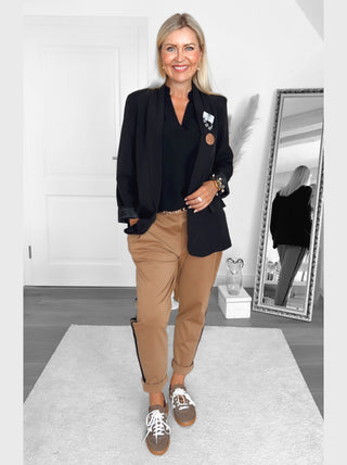 Frau in heller, minimalistischer Umgebung trägt die OFF#DLY Pamt Turin camel, camel-farbene Hose mit hochgekrempeltem Saum, kombiniert mit schwarzem Blazer, betont durch sportliche seitliche Streifen für dynamischen Chic.
