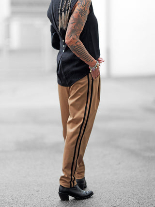 Person trägt OFF#DLY Pamt Turin camel Hose mit schwarzen Seitenstreifen, kombiniert mit schwarzen Stiefeln. Die sportlich-chice Hose hat einen konischen Schnitt und elastischen Bund, ideal für Komfort.