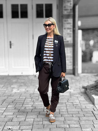 Frau trägt OFF#DLY Pamt Turin brown, eine sportlich-schicke Hose mit elastischem Bund und schwarzen Seitenstreifen, kombiniert mit Sneakers und schwarzem Blazer.