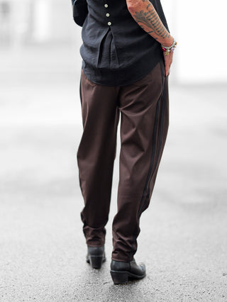 Person trägt die OFF#DLY Pamt Turin brown, eine glänzende, braune Hose mit schwarzem Seitenstreifen, kombiniert mit schwarzen Lederstiefeletten. Tattoos und Perlenarmbänder sind am Arm sichtbar.